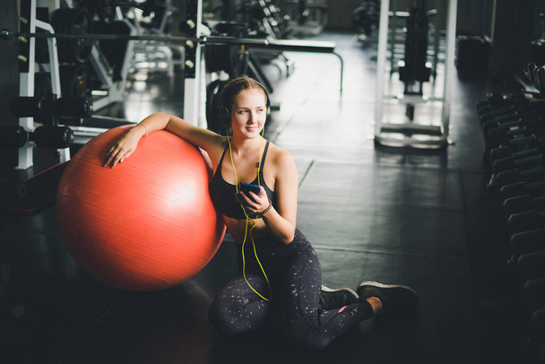 A beautiful woman wearing a sports shirt is sitting and relaxing by listening to music from headphones. With rubber  yoga ball or exercise ball - Foto, immagini