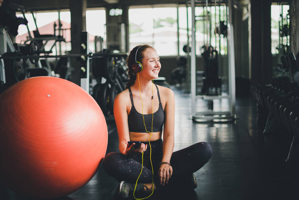 A beautiful woman wearing a sports shirt is sitting and relaxing by listening to music from headphones. With rubber  yoga ball or exercise ball - Foto, Imagem