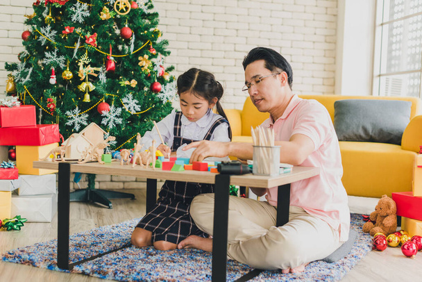 Азиатский отец и дочь играют в игрушки на столе в гостиной украшенные елками для подготовки к празднованию Рождества и Нового года
. - Фото, изображение