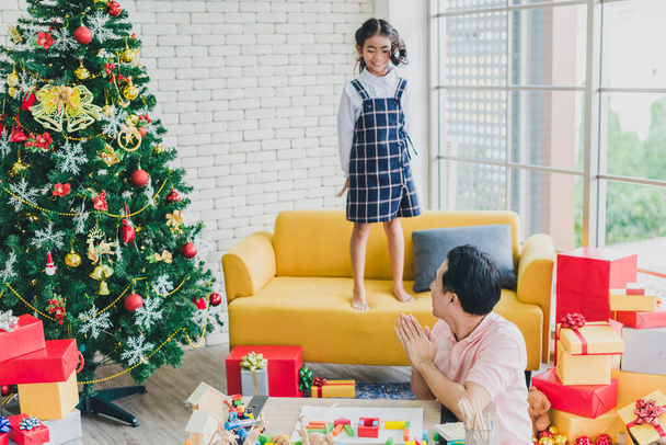Azjatycki ojciec bawi się z córką skacząc z sofy w salonie ozdobionym choinkami, aby przygotować się do obchodów Bożego Narodzenia i Nowego Roku. - Zdjęcie, obraz