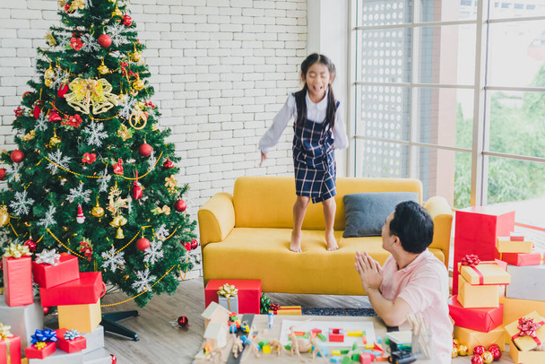 Père asiatique joue avec sa fille sautant du canapé dans le salon décoré avec des arbres de Noël pour préparer la célébration de Noël et du Nouvel An
. - Photo, image