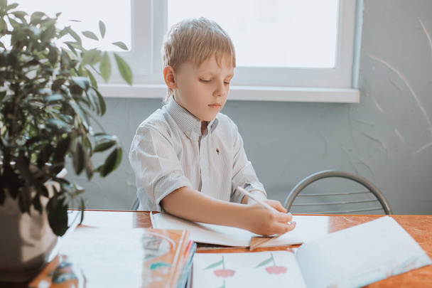 El niño escribe en un cuaderno en casa durante la cuarentena CoVid-19, a distancia de aprendizaje en línea con un ordenador portátil, un niño haciendo los deberes para la escuela. Los niños se quedaron en casa
. - Foto, imagen