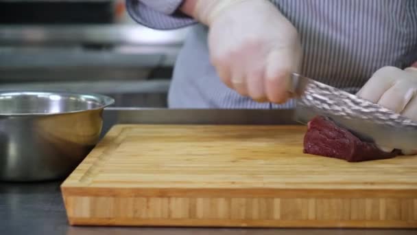 Chef cook in kitchen takes large fillet venison from an iron bowl and with knife cuts meat into pieces for dish, on wooden bamboo Board. Working as cook in kitchen of restaurant, preparing for dish. - Кадры, видео
