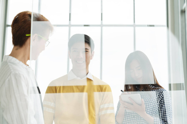 Group of young asian attractive business people standing, talking and listening to meeting with manager behind transparent glass wall. Brainstorming ideas with team in casual business meeting concept. - Фото, изображение