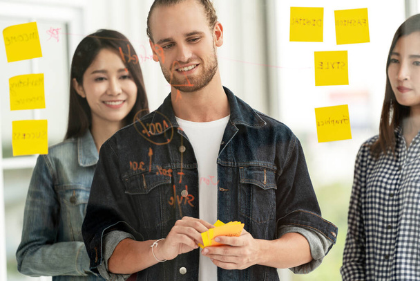 Groep van jonge succesvolle creatieve multi-etnische team glimlach en brainstorm op project op kantoor met gele postnota of plakkerige nota. Blanke man kijkt naar glazen muur concentreren op het delen van idee. - Foto, afbeelding