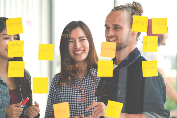 Hipster attractive young caucasian man and young asian chinese girl talk and brainstorming together at glass wall sharing business ideas project plans with post it sticky note. Marketing plan concept. - Photo, Image