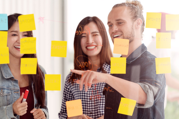 Group of young successful creative multiethnic team smile and brainstorm on project together in modern office with post note or sticky note. Caucasian man point on glass wall sharing idea. - Photo, Image