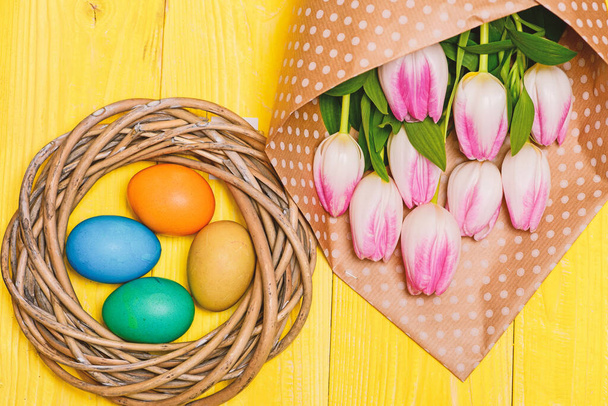Jarní prázdniny. Veselé Velikonoce. Blíží se Velikonoce. Sbírám velikonoční vajíčka. Jarní vibrace. Barevná vejce a kytice čerstvé tulipán květiny na žlutém pozadí horní pohled. Tradice slaví Velikonoce - Fotografie, Obrázek