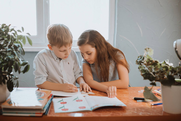 Το αγόρι και το κορίτσι διαβάζοντας το βιβλίο στο σπίτι κατά τη διάρκεια της καραντίνας CoVid-19, εξ αποστάσεως μάθηση σε απευθείας σύνδεση με ένα φορητό υπολογιστή, ένα παιδί κάνει την εργασία για το σχολείο. Τα παιδιά έμειναν σπίτι.. - Φωτογραφία, εικόνα