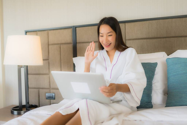 Portré gyönyörű fiatal ázsiai nők segítségével laptop vagy számítógép mobiltelefon az ágyon a hálószoba belső - Fotó, kép