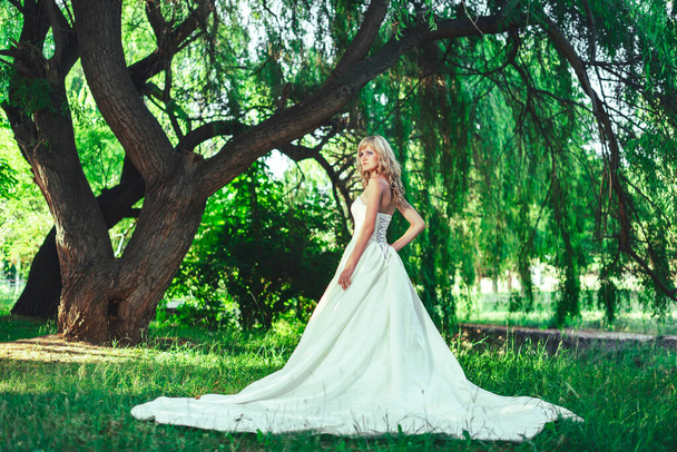 Dziewczyna w sukni ślubnej z pociągiem w ogrodzie Park. Kobieta w białej sukni wieczorowej w lesie. Panna młoda w długiej sukni na tle zielonych drzew - Zdjęcie, obraz
