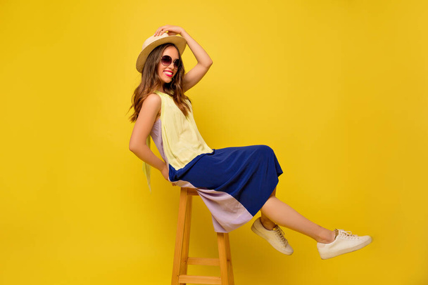 Ritratto a figura intera di felice ragazza uscita indossando giallo brillante con abito blu, occhiali da sole e cappello rilassante sulla sedia con sorriso gentile
 .  - Foto, immagini