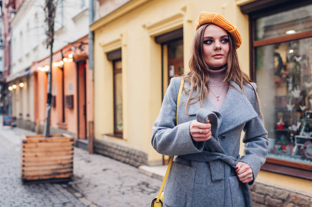 Portrait of stylish young woman wearing yellow knitted beret grey tying coat holding purse on street outdoors. Spring fashion female accessories and clothes. Trendy classic retro outfit - Photo, Image