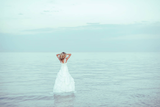 Ein Mädchen steht im weißen Hochzeitskleid im Wasser. Eine Frau im weißen Kleid am Ufer des Issyk-Kul-Sees in Kirgisistan. Braut in der Nähe des Sees - Foto, Bild
