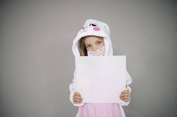 beautiful girl in easter bunny costume and face protective mask holding white board sign, concept of easter during coronavirus lockdown  - Фото, изображение