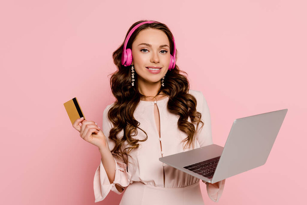 mujer joven feliz en auriculares inalámbricos utilizando el ordenador portátil y la celebración de la tarjeta de crédito aislado en rosa
  - Foto, imagen