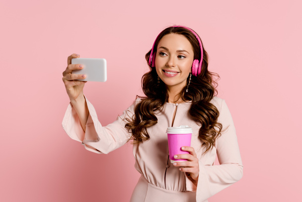 happy girl in wireless headphones taking selfie and holding paper cup on pink  - Photo, Image