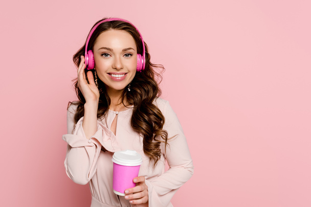 χαρούμενο κορίτσι ακούει μουσική σε ασύρματα ακουστικά και κρατώντας χάρτινο κύπελλο απομονωμένο σε ροζ  - Φωτογραφία, εικόνα