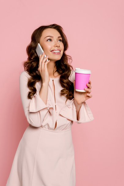 χαρούμενο κορίτσι μιλάει στο smartphone και κρατώντας χάρτινο κύπελλο απομονωμένο σε ροζ  - Φωτογραφία, εικόνα