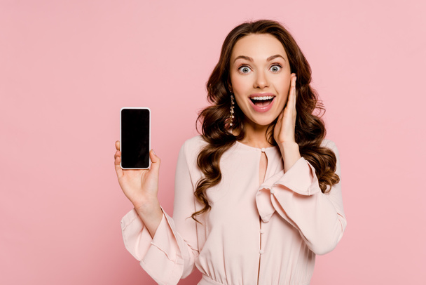 vzrušená dívka drží smartphone s prázdnou obrazovkou a dívá se na kameru izolované na růžové  - Fotografie, Obrázek