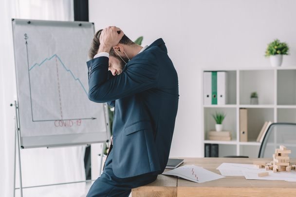 боковой вид депрессивного бизнесмена, держащего руки на кипящей голове, находясь рядом с флипчартом и рабочим местом в офисе
 - Фото, изображение