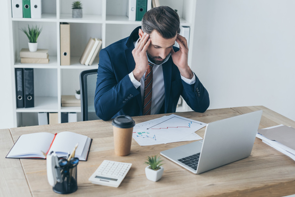 estressado, homem de negócios exausto tocando a cabeça enquanto sentado com os olhos fechados no local de trabalho perto de documentos e laptop
  - Foto, Imagem