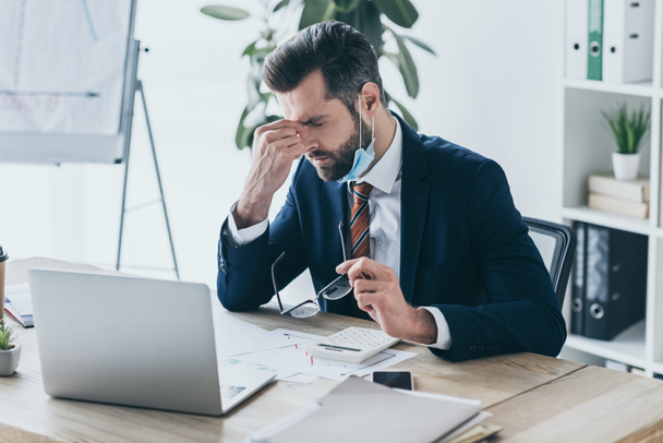 καταθλιπτικός, εξαντλημένος επιχειρηματίας κρατώντας γυαλιά και αγγίζοντας το πρόσωπο ενώ κάθεται στο χώρο εργασίας κοντά στο laptop και τα έγγραφα - Φωτογραφία, εικόνα