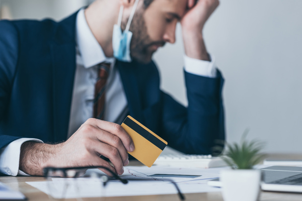 selektivní zaměření rozčileného podnikatele držícího kreditní kartu při sezení na pracovišti se zavřenýma očima a skloněnou hlavou - Fotografie, Obrázek