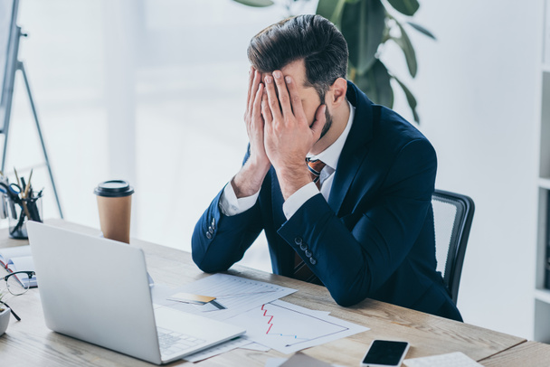 depressed businessman covering face with hands while sitting at workplace near laptop - Valokuva, kuva
