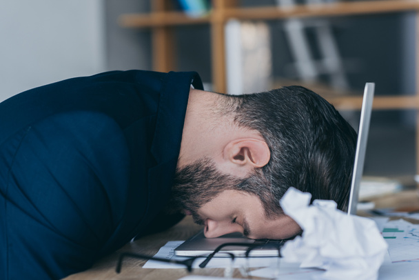 hombre de negocios deprimido con los ojos cerrados sentado en el lugar de trabajo con la cabeza en el ordenador portátil cerca de papel arrugado
 - Foto, Imagen