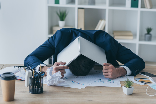 депресивний бізнесмен прикриває голову ноутбуком, сидячи на робочому місці
 - Фото, зображення
