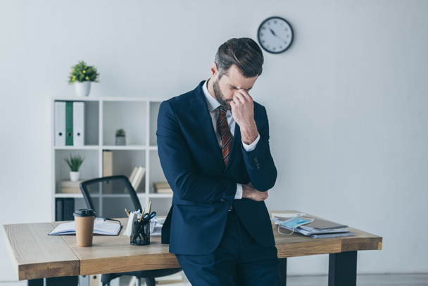 hombre de negocios deprimido con los ojos inclinados y cerrados tocando la cara mientras está de pie en el lugar de trabajo
  - Foto, Imagen