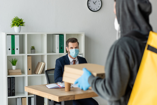 selective focus of food delivery man holding pizza boxes near businessman in medical mask sitting at workplace in office - Foto, afbeelding