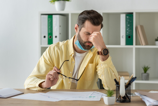 виснажений бізнесмен тримає окуляри і торкається обличчя, сидячи на робочому місці біля документів
 - Фото, зображення