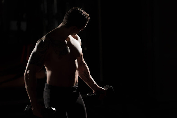 Handsome power athletic guy bodybuilder doing exercises with dumbbell. Fitness muscular body on dark background. - Foto, Bild