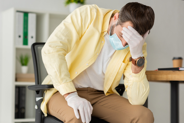 depressed businessman in latex gloves and medical mask holding hand on forehead while sitting in office - Photo, Image
