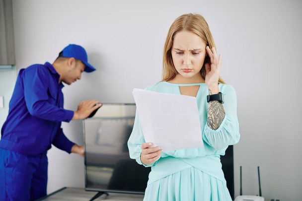 Frowning unhappy young woman reading document with price of tv set installation - Foto, Imagem