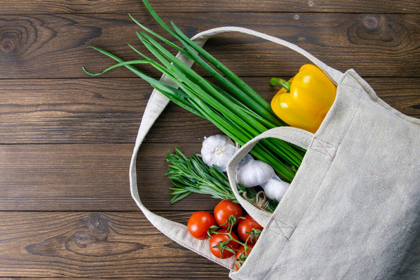 Υφαντή τσάντα από διαφορετικά τρόφιμα για την υγεία σε σκούρο ξύλινο τραπέζι φόντο. Πράσινο κρεμμύδι, σκόρδο, ντομάτα και κίτρινη πιπεριά. Κάτοψη με ελεύθερο χώρο. - Φωτογραφία, εικόνα