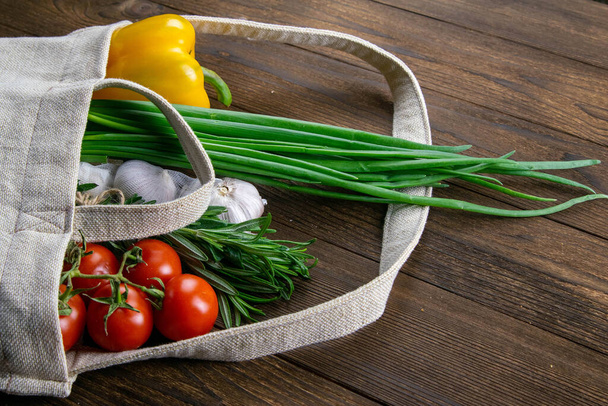 Koyu ahşap masa arkasında farklı sağlıklı yiyeceklerden örülmüş bir çanta. Yeşil soğan, sarımsak, domates ve sarı biber. Açık alanlı en üstteki görünüm. - Fotoğraf, Görsel