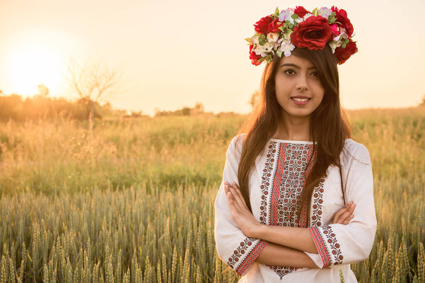 Sárga tónusú portré arany órában. Csinos fiatal nő visel hagyományos ukrán ruhát és virágkoszorú séta a búzamezőn, gyönyörű etnikai lány kézzel készített díszített virágos korona csodálja a természetet.  - Fotó, kép