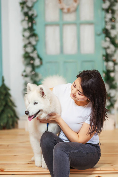 Lány egy kutya Samoyed laika a háttérben a karácsonyi dekorációk. Egy nő fehér kutyával a karácsonyi tornácon. Ázsiai nő fehér husky - Fotó, kép