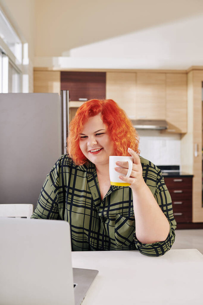 Smiling overweight young woman with red hair drinking morning coffee in kitchen and checking e-mail on her laptop - Zdjęcie, obraz