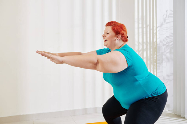 Happy young overweight woman in sportswear enjoying doing squats at home, training legs, trying to form perfect butt shape - Foto, imagen