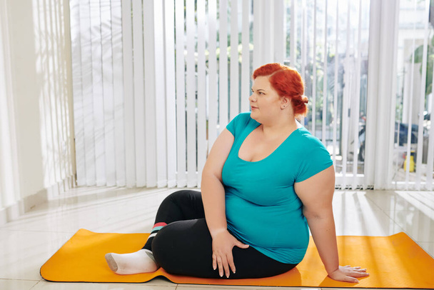 Túlsúlyos fiatal nő ül jóga szőnyegen, és pihenés edzés után a fitneszklubban - Fotó, kép