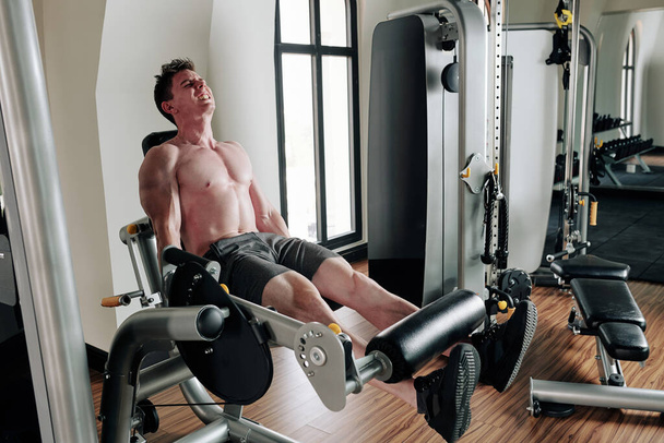 Jonge sporter bijna schreeuwen bij het doen van beenverlenging met zwaar gewicht - Foto, afbeelding