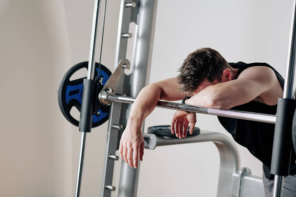 Spor salonunda ağırlık kaldırdıktan sonra halterlere yaslanan yorgun genç vücut geliştirici. - Fotoğraf, Görsel