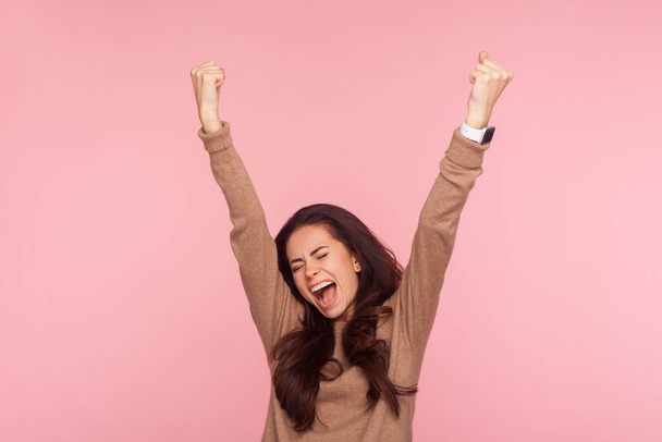 I am champion! Portrait of overjoyed enthusiastic young woman with brunette hair raising her hands and screaming ecstatically about victory, enjoying triumph. studio shot isolated on pink background - Foto, immagini