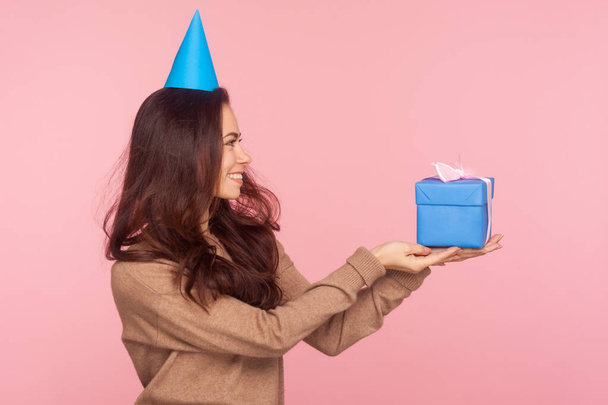 Take this present! Side view of happy generous nice young woman wearing party cone on head holding gift box, congratulating and offering birthday present. studio shot isolated on pink background - Foto, imagen