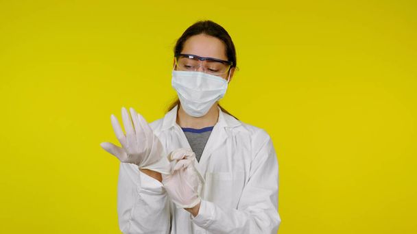 Νεαρός γιατρός με ιατρική μάσκα και γυαλιά ασφαλείας βάζει λατέξ γάντια στα χέρια του - Φωτογραφία, εικόνα