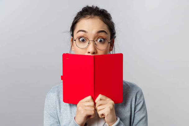 Education, university and people concept. Close-up portrait of surprised and amused young asian girl, widen eyes at amazing interesting story, cover mouth behind red planner, stand grey background - 写真・画像